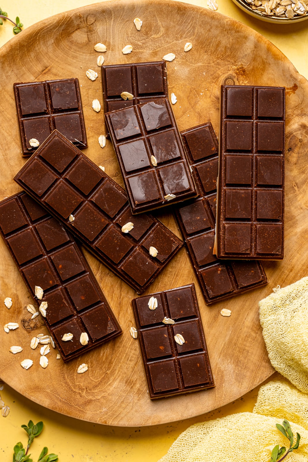 Up close shot of chocolate bars on a brown wood plate with dry oats on a yellow table with a yellow napkin.