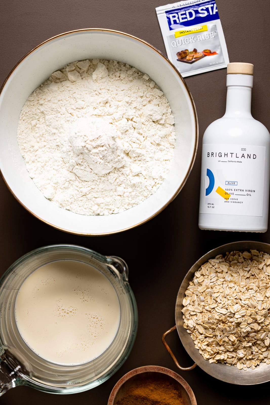 Ingredients on a brown table including flour, olive oil, yeast, milk, oats, and cinnamon.