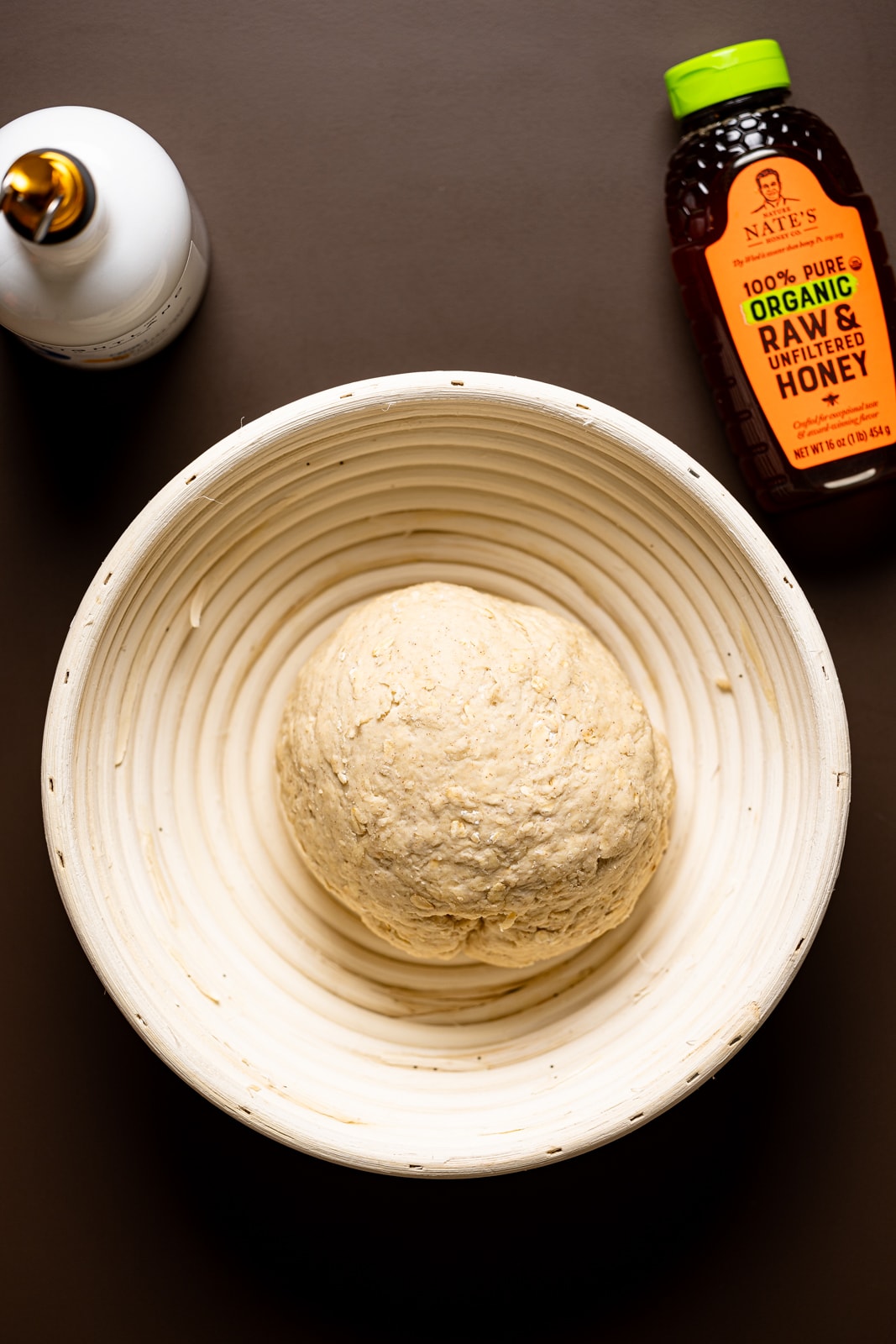 Bread dough in a bread proofing bowl with honey and olive oil on a brown table.