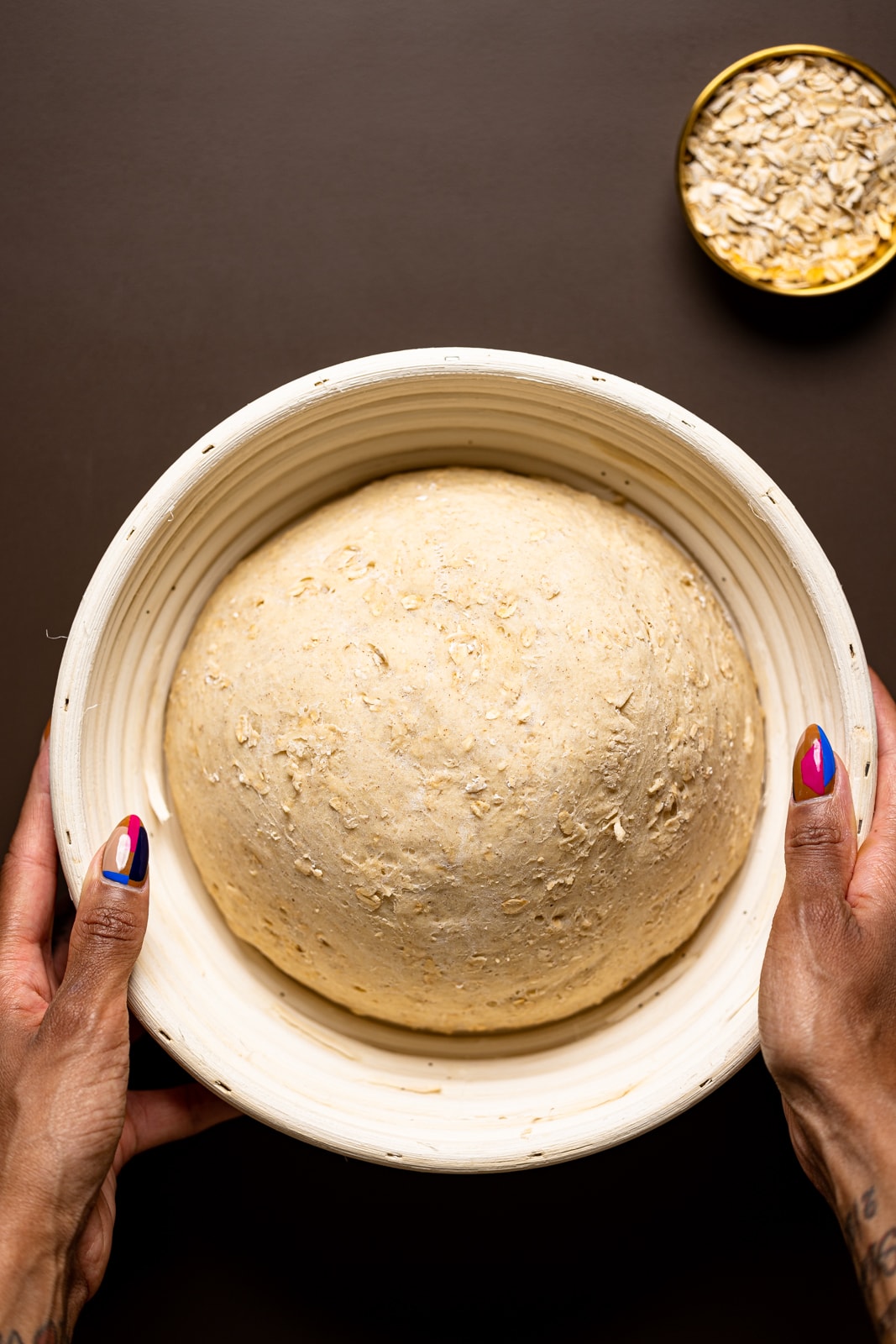Risen bread dough in a bread proofing bowl being held on a brown table.