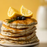 Stack of Vegan Lemon Poppyseed Pancakes topped with lemon wedges, blueberries, and a sprig of rosemary