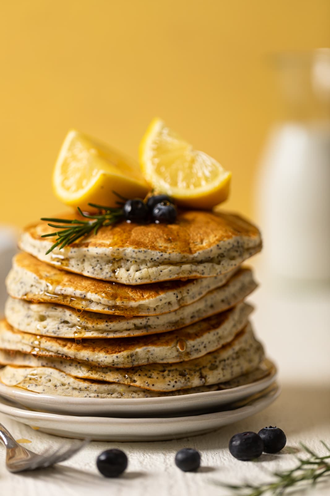 Stack of Vegan Lemon Poppyseed Pancakes topped with lemon wedges, blueberries, and a sprig of rosemary