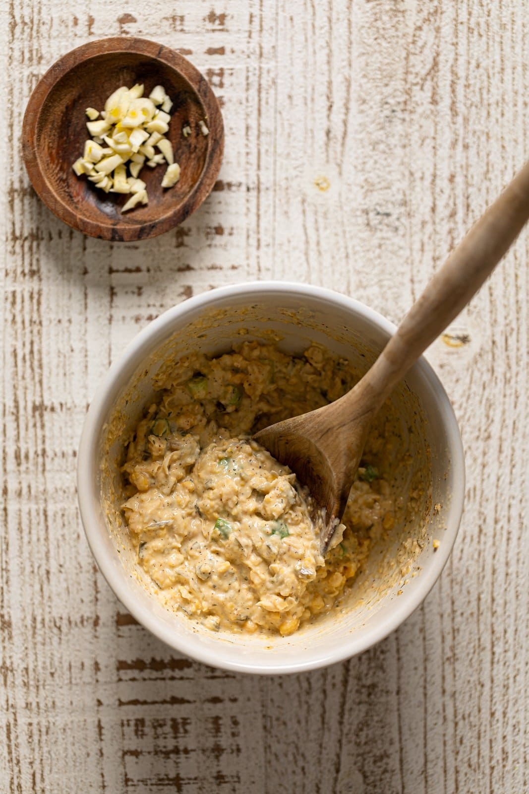 Wooden spoon stirring a bowl of vegan \"tuna\" near a bowl of garlic