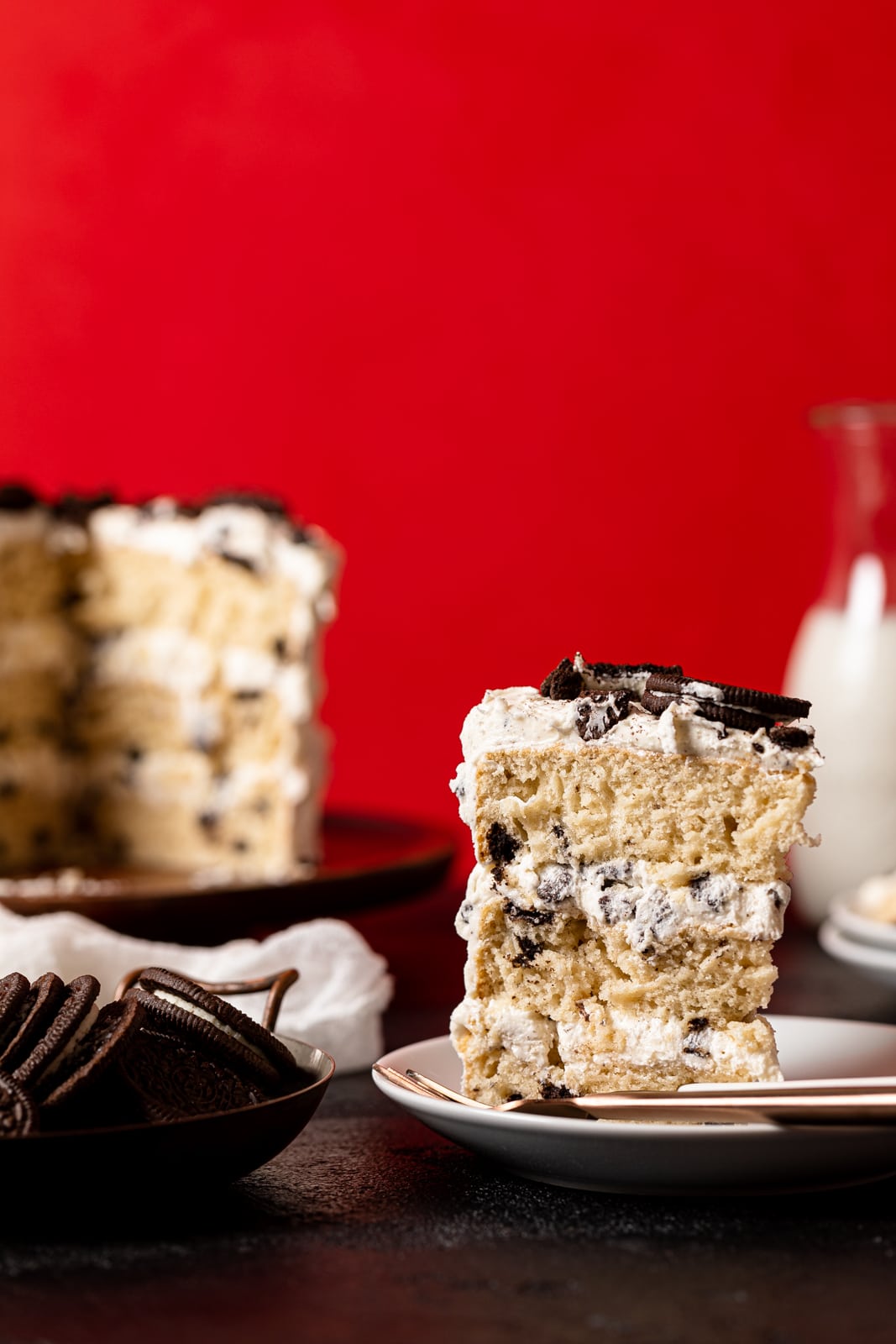 A slice of Oreo cake standing on a white plate with a fork and the whole cake on the cake stand in the background with a bowl of Oreo cookies on a black table and red background. 