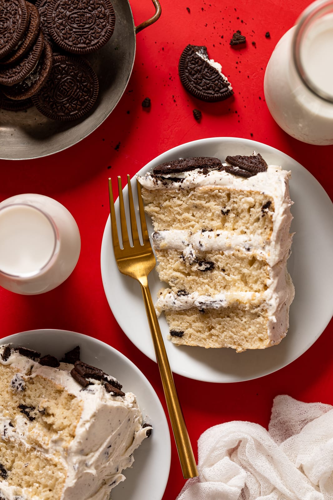 Three Vanilla oreo cake slices on white plates with two forms, a glass of milk, and Oreo cookie pieces on a red table.