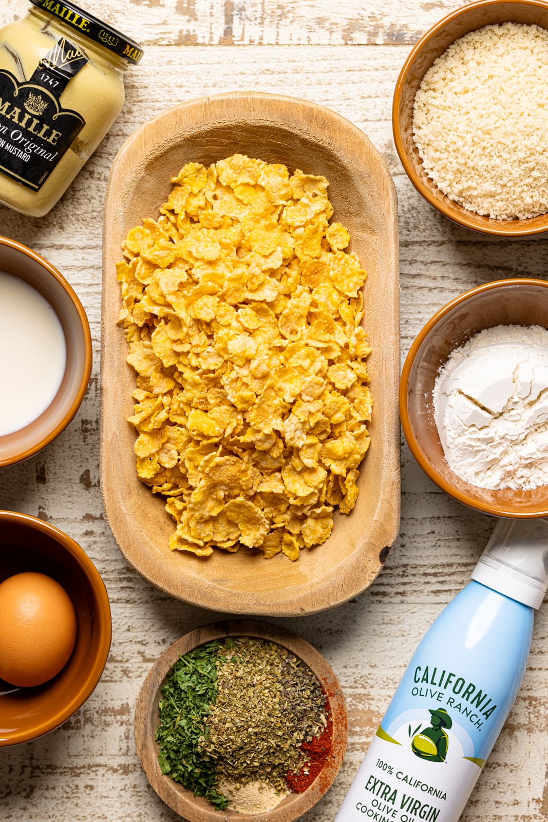 Ingredients on a white wood table including cornflakes, oil spray, eggs, breadcrumbs, flour, dijon mustard, and milk.