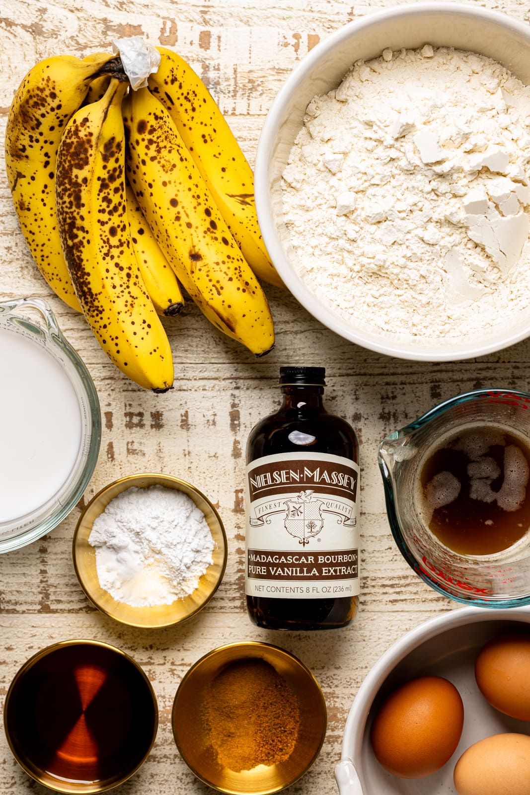 Ingredients on a white wood table including banana, flour, vanilla, eggs, milk, and maple syrup.