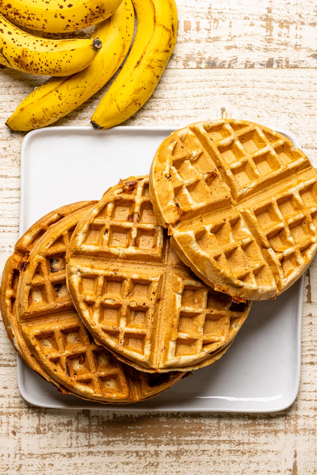 Waffles on a white plate on a white wooden table with bananas.