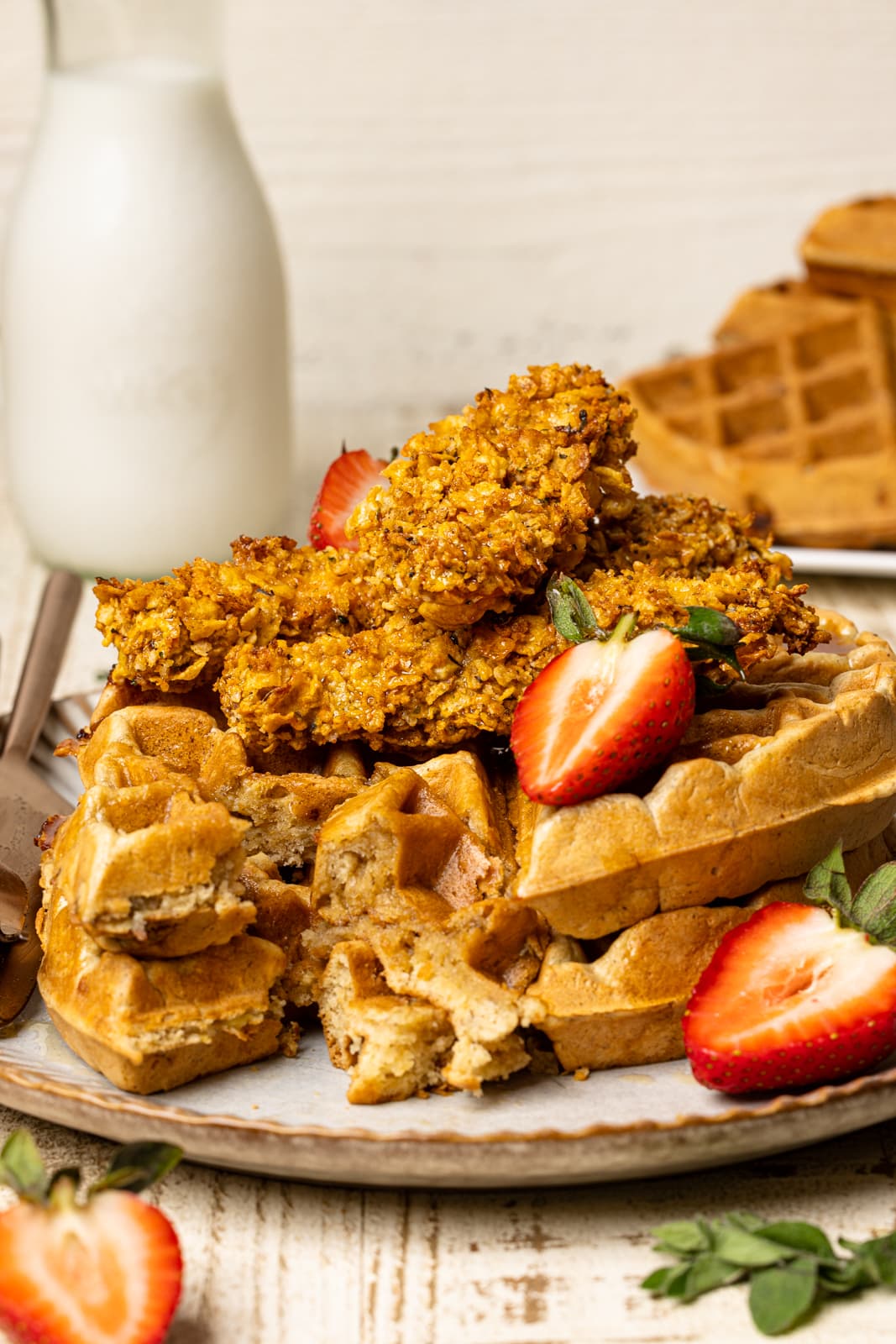 Chicken and waffles on a white plate on a white wood table with a glass of milk and strawberries.