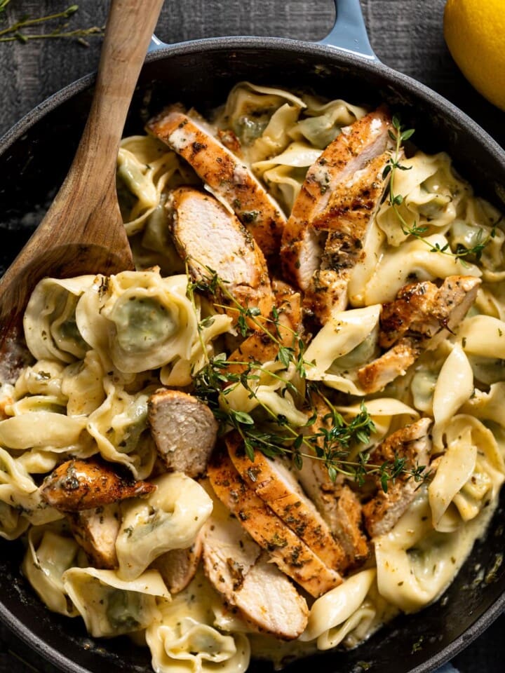 Wooden spoon stirring a skillet of Creamy Lemon Herb Roasted Chicken with Spinach Tortellini