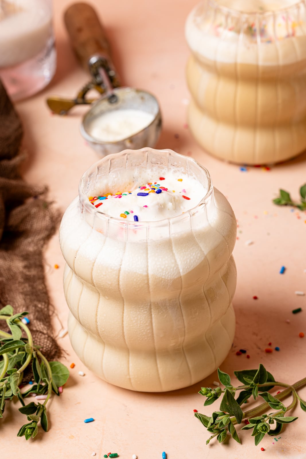 Two glasses of latte on a peach table with herbs, sprinkles, and an ice cream sccop.