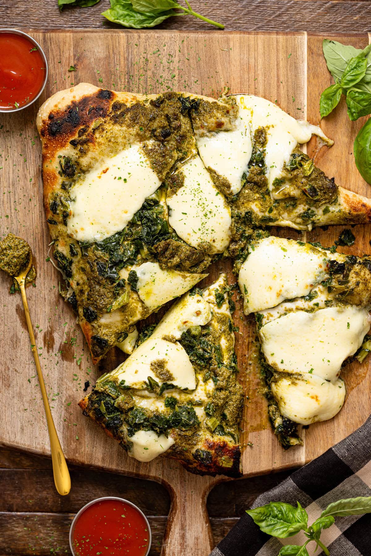 Pizza sliced on a cutting board with a spoon, sauces, and basil leaves.