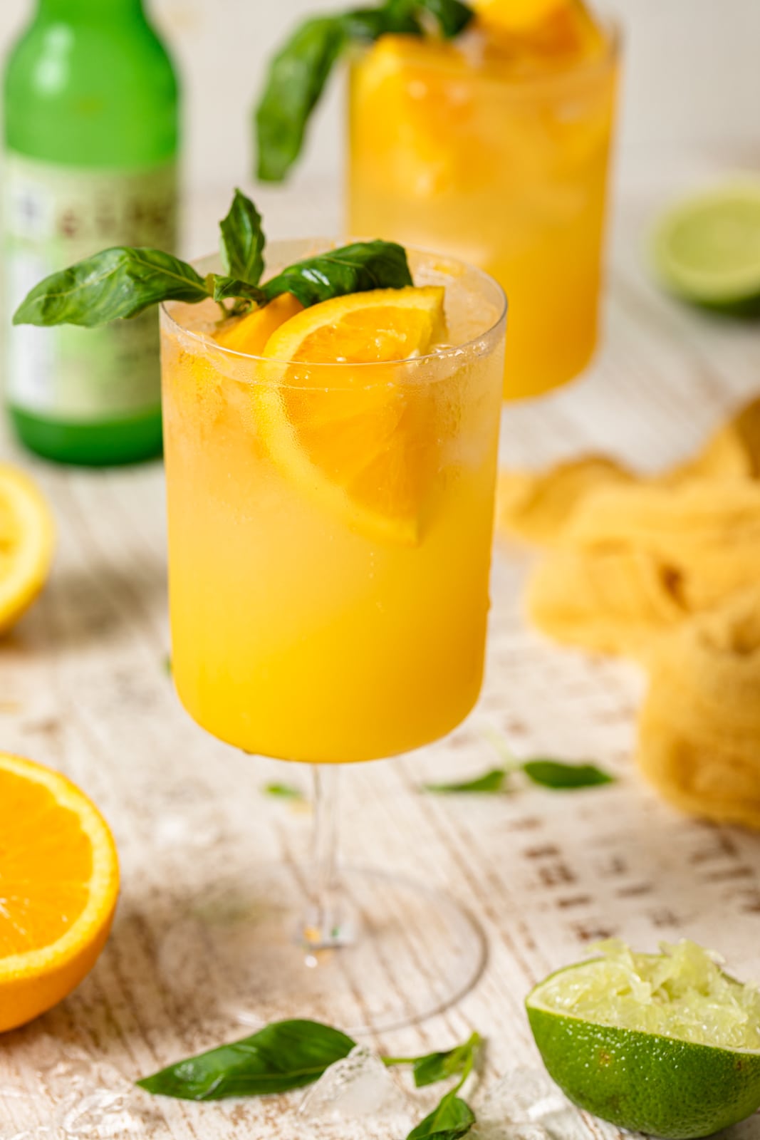 Bright Orange Crush Mocktail with Basil on a table with oranges, limes, basil leaves, ice, and other ingredients