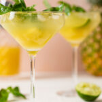 Two long-stemmed glasses of Pineapple Coconut Basil Mocktail with Lime topped with basil leaves