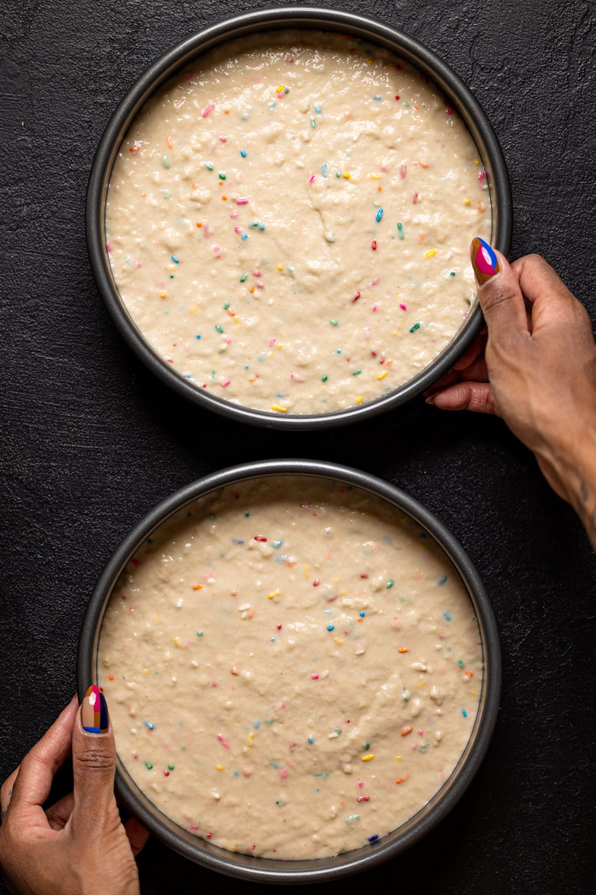 Hands holding two round pans filled with vegan funfetti cake batter
