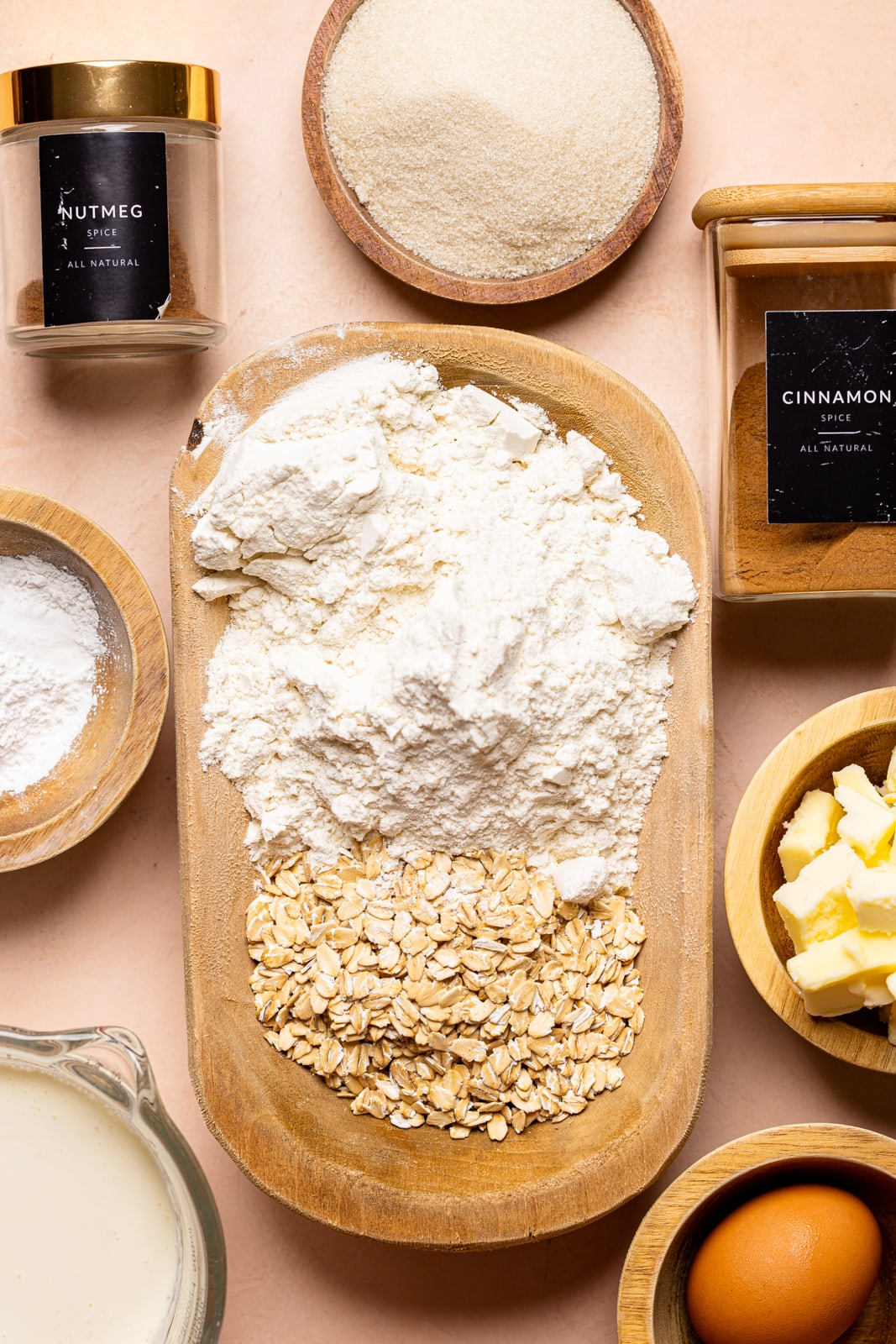 Ingredients on a peach table including flour, oats, sugar, spices, egg, milk, butter, and baking powder.