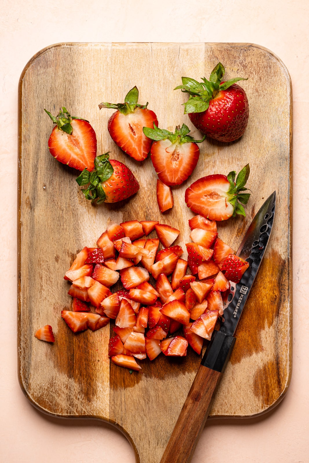 Diced and sliced strawberries on a wood cutting board with a knife on a peach table.