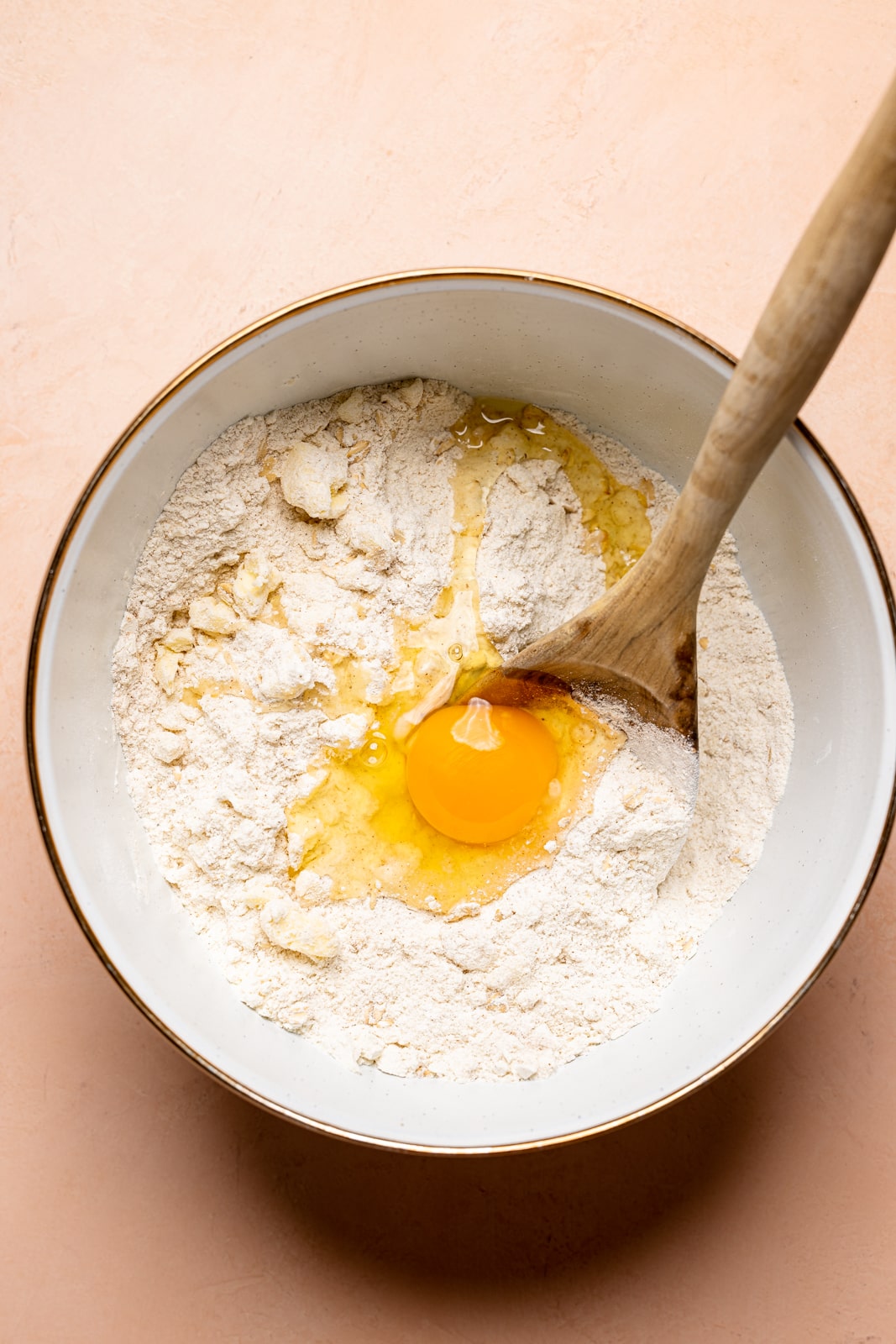 Dry ingredients in a large white bowl with eggs and a wooden spoon on a peach table.