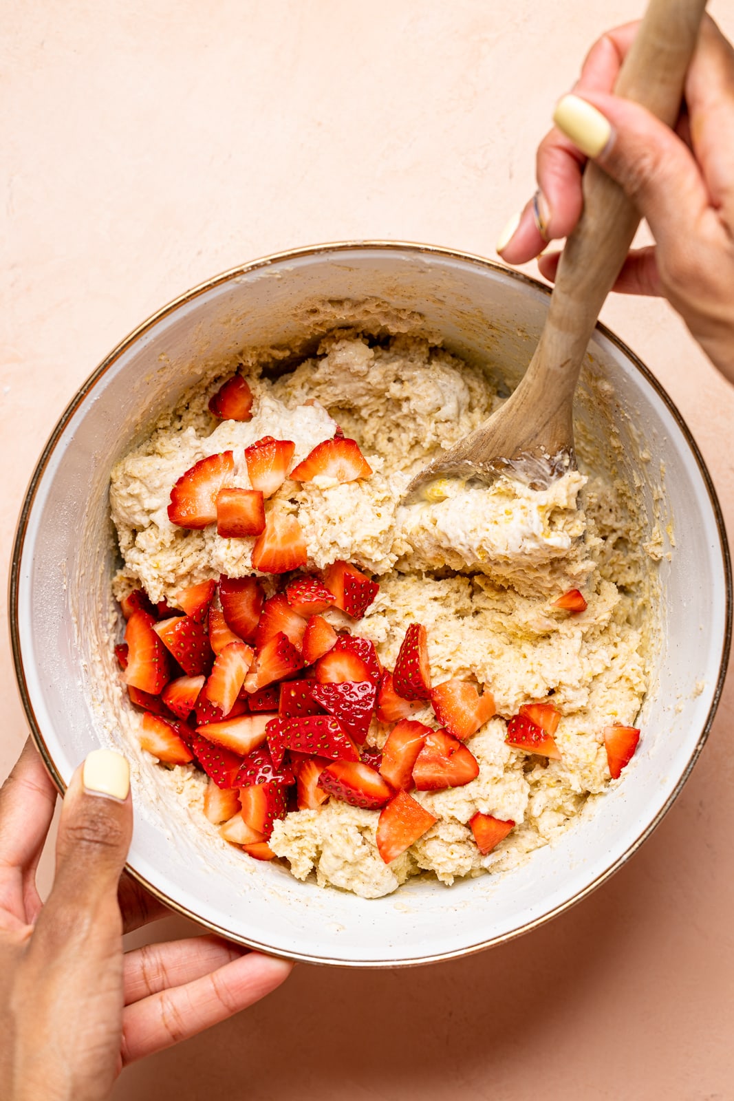 Scones batter in a large white bowl with diced strawberries and a wooden spoon being stirred on a peach table.