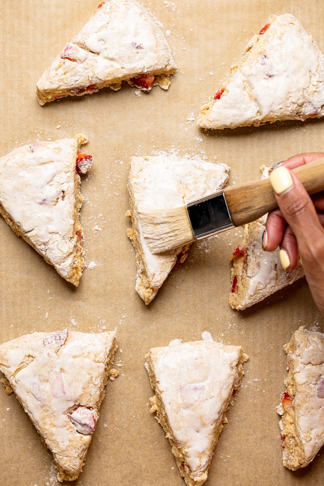 Triangle shaped scones on a baking sheet with parchment paper being brushed with egg wash.