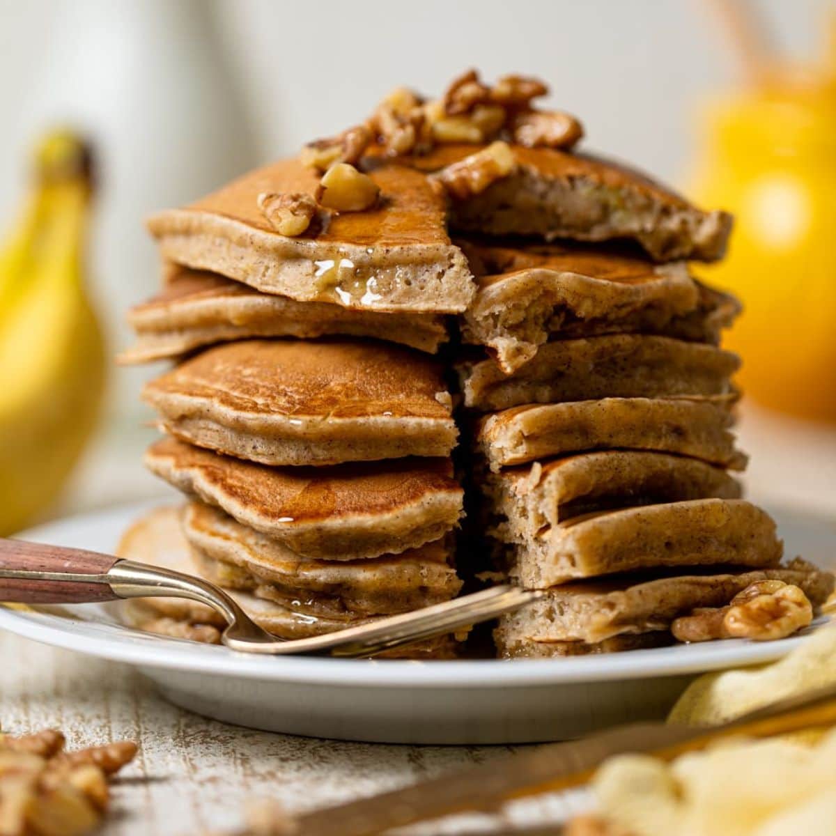 Stack of pancakes on a white plate sliced with a fork and bananas in the background.