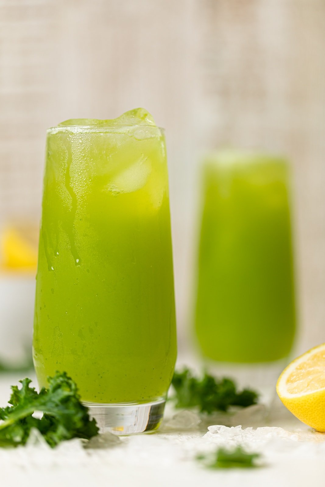 Two glasses of bright green Kale Lemonade next to kale, lemons, and ice