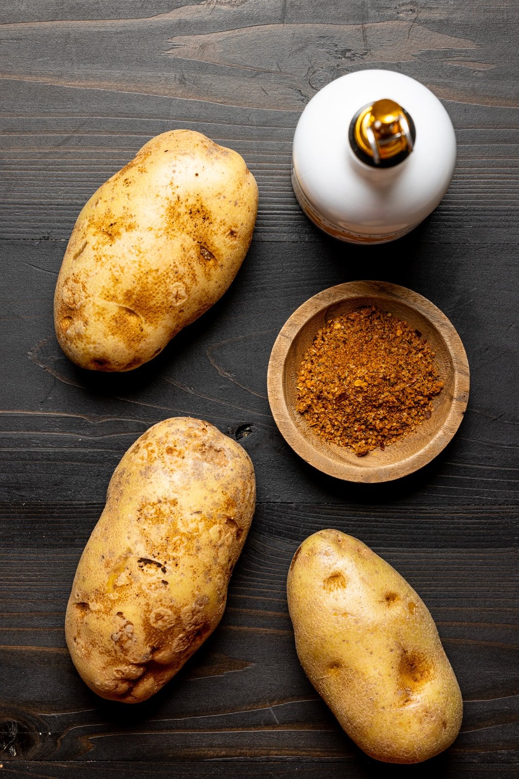 Three large russet potatoes on a black wood table with Cajun seasoning, and a bottle of olive oil.