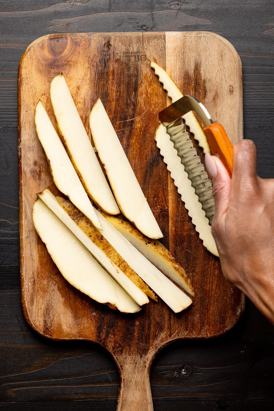 Russet potatoes being cut with crinkle cutter tool on a cutting board on black wood table.
