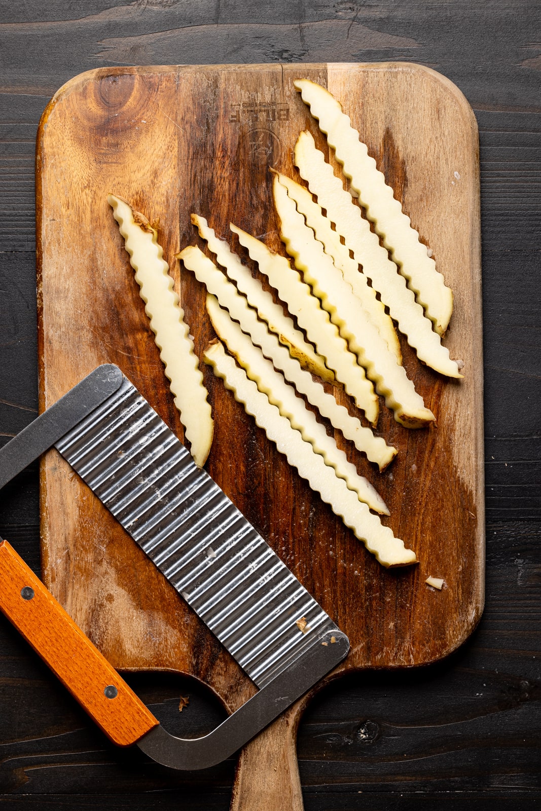 Fries cut into crinkled shape using crinkle cut tool on a cutting board on a black wood table.