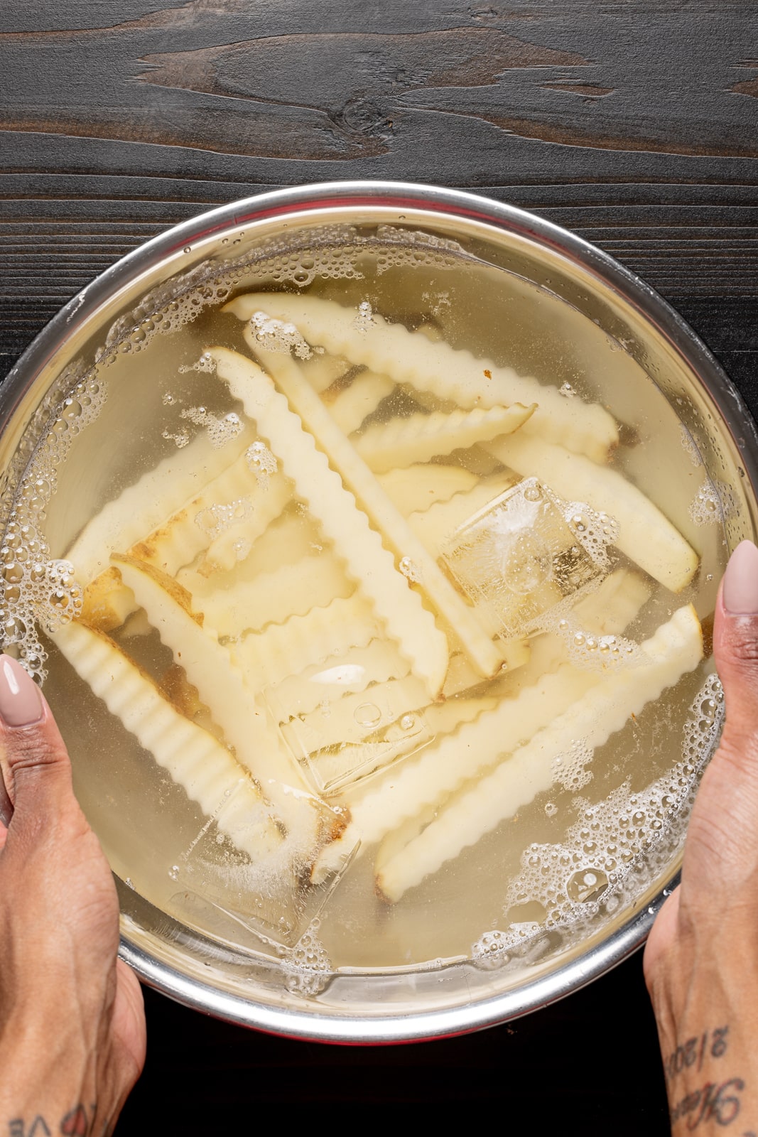 Fries soaking in cold water in a silver bowl on a black wood table.