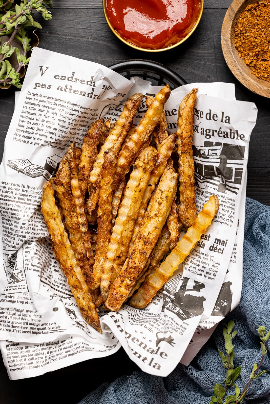 Fries in a food basket on black wood table with ketchup, seasoning, and a blue napkin.