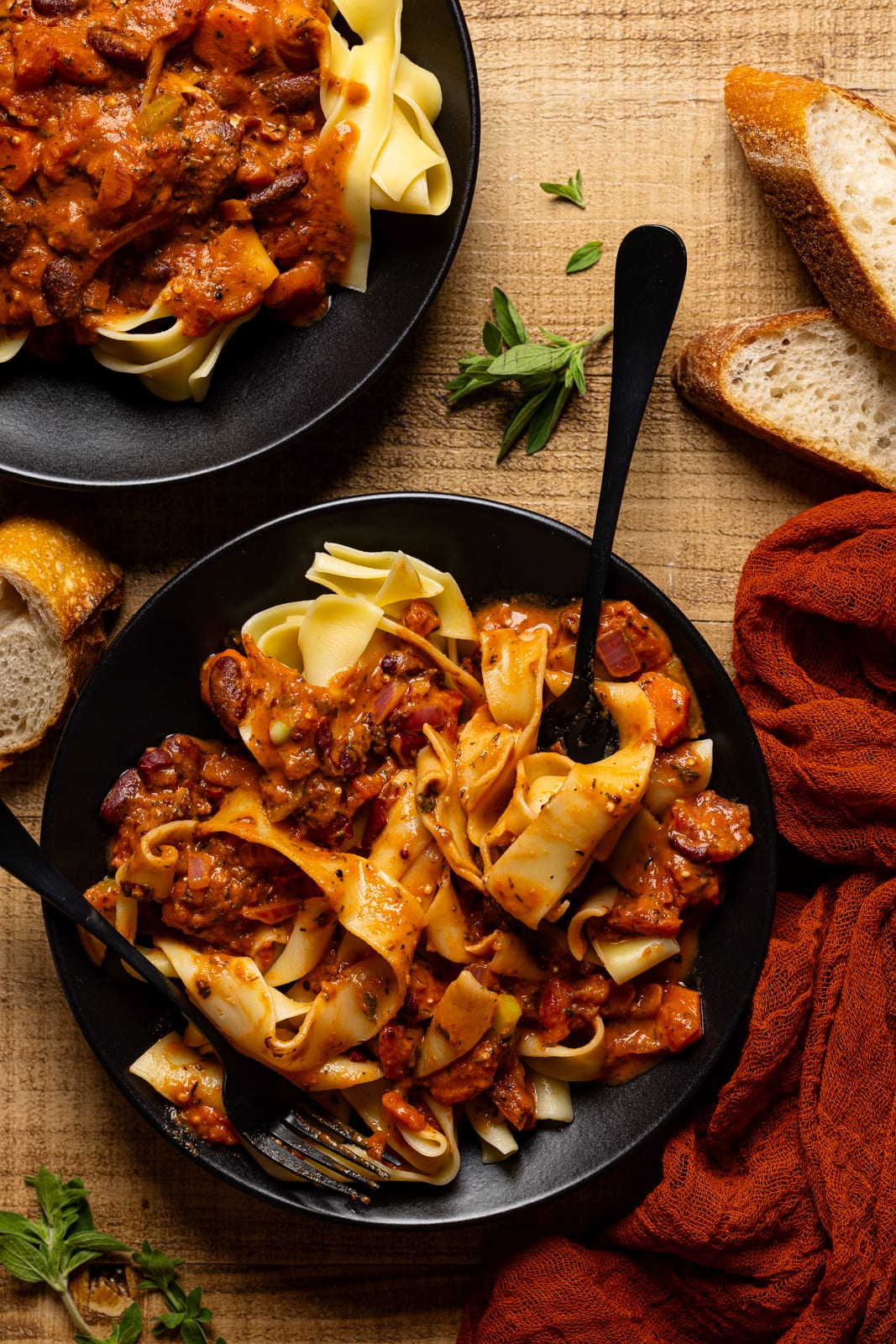 Two plates of pasta with two black forks and bread on the side.