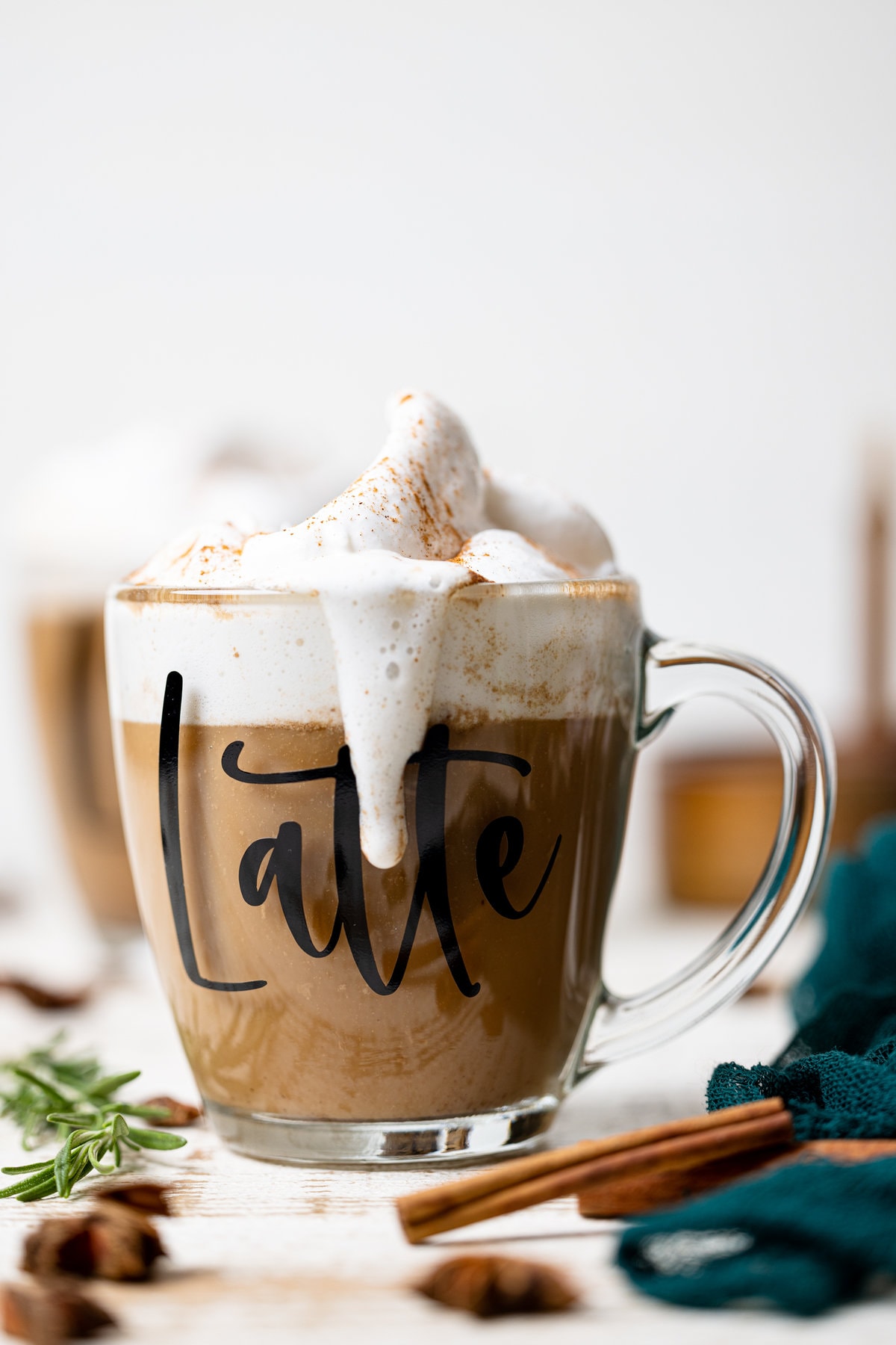 Overflowing glass of Fall Spice Latte with the word \"Latte\" on the side near cinnamon sticks and a sprig of rosemary