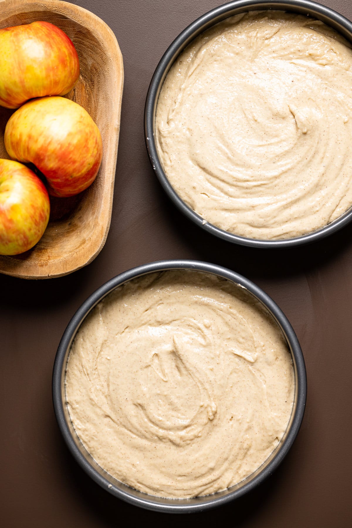 Two circle pans filled with Apple Cider Cake batter next to a bowl of apples