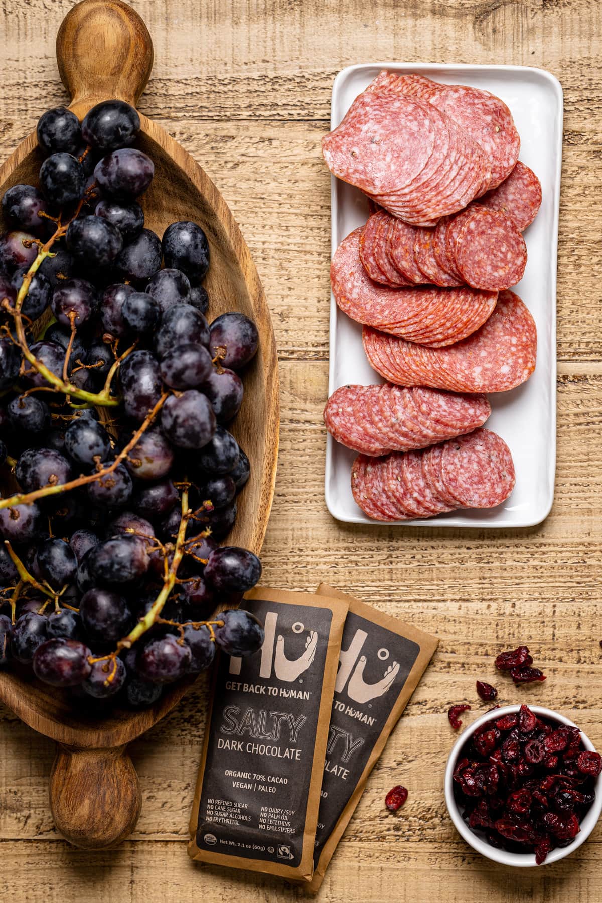 Wooden bowl of black grapes next to a plate of cured meats, bowl of dried cranberries, and Get Back to Human salty dark chocolate