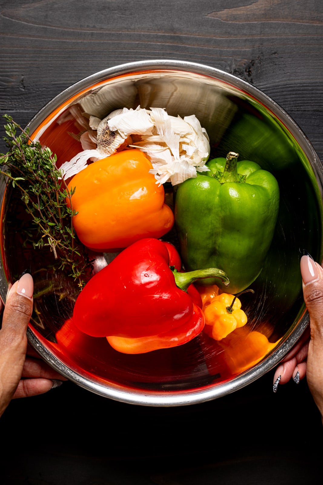Silver bowl being held full of bell peppers, garlic, thyme, and peppers.