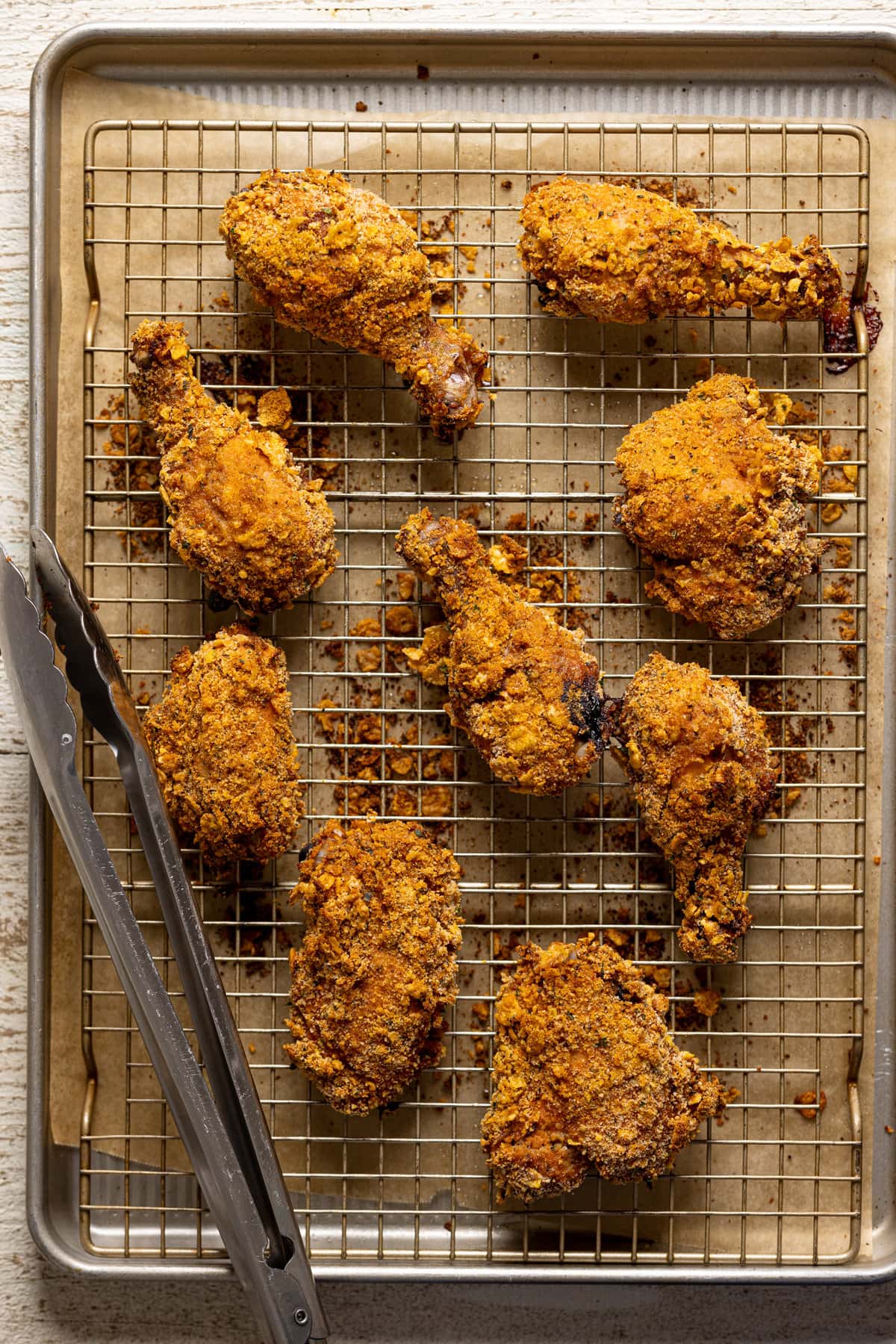 Baked fried chicken on a wire rack on a baking sheet lined with parchment paper with silver tongs.