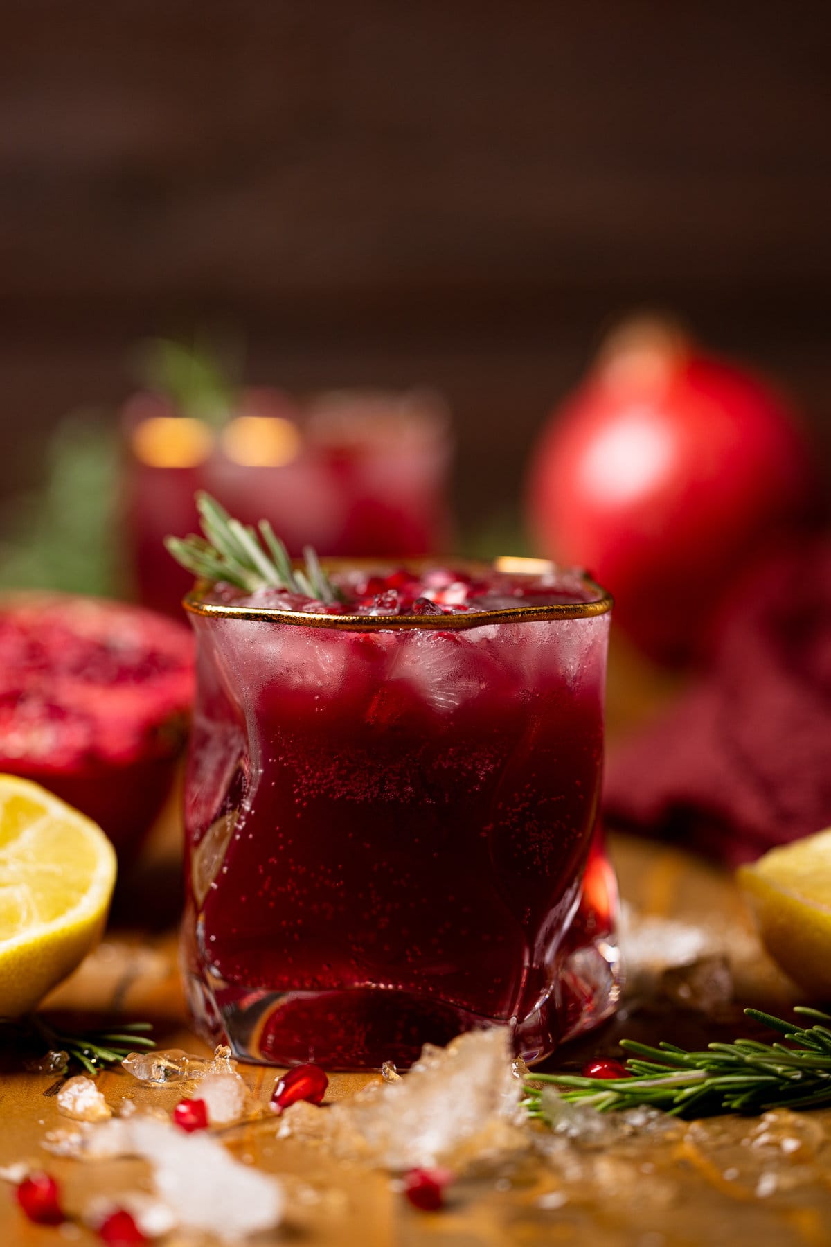 Holiday Pomegranate Ginger Mocktail in a gold-rimmed glass garnished with rosemary and pomegranate seeds near lemon and ice