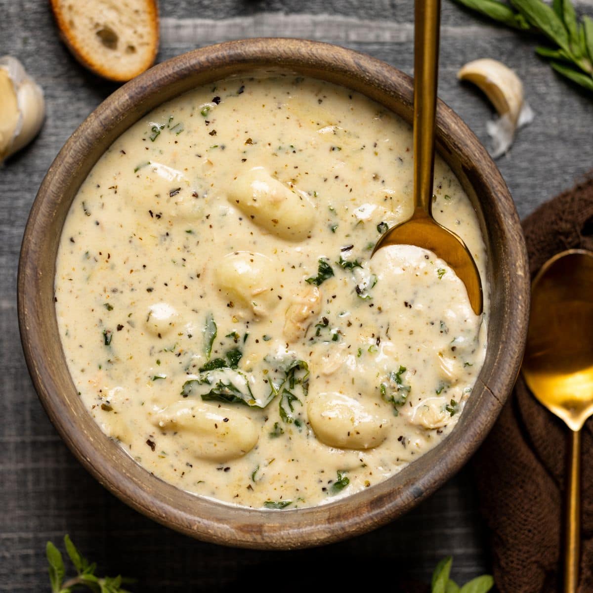 Up close shot of creamy soup in a brown wood bowl on a grey wood table with two gold spoon and side of bread.