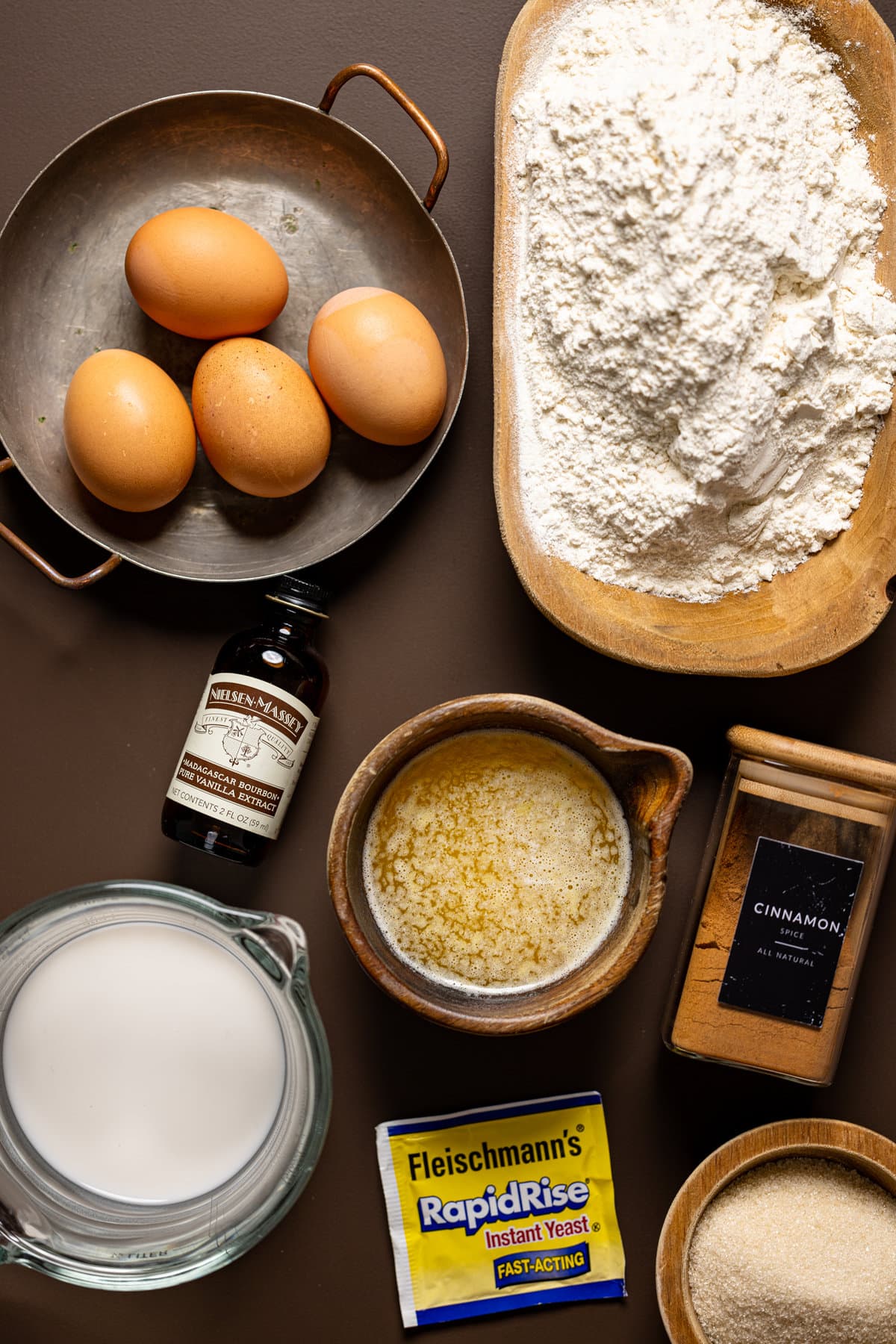 Ingredients for doughnuts which include flour, eggs, vanilla, butter, cinnamon, instant yeast, sugar, and milk on a dark brown table. 