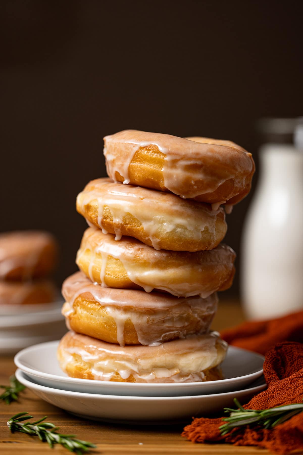 Glazed doughnuts stacked tall on two white plates on a brown wood table with milk in the background. 