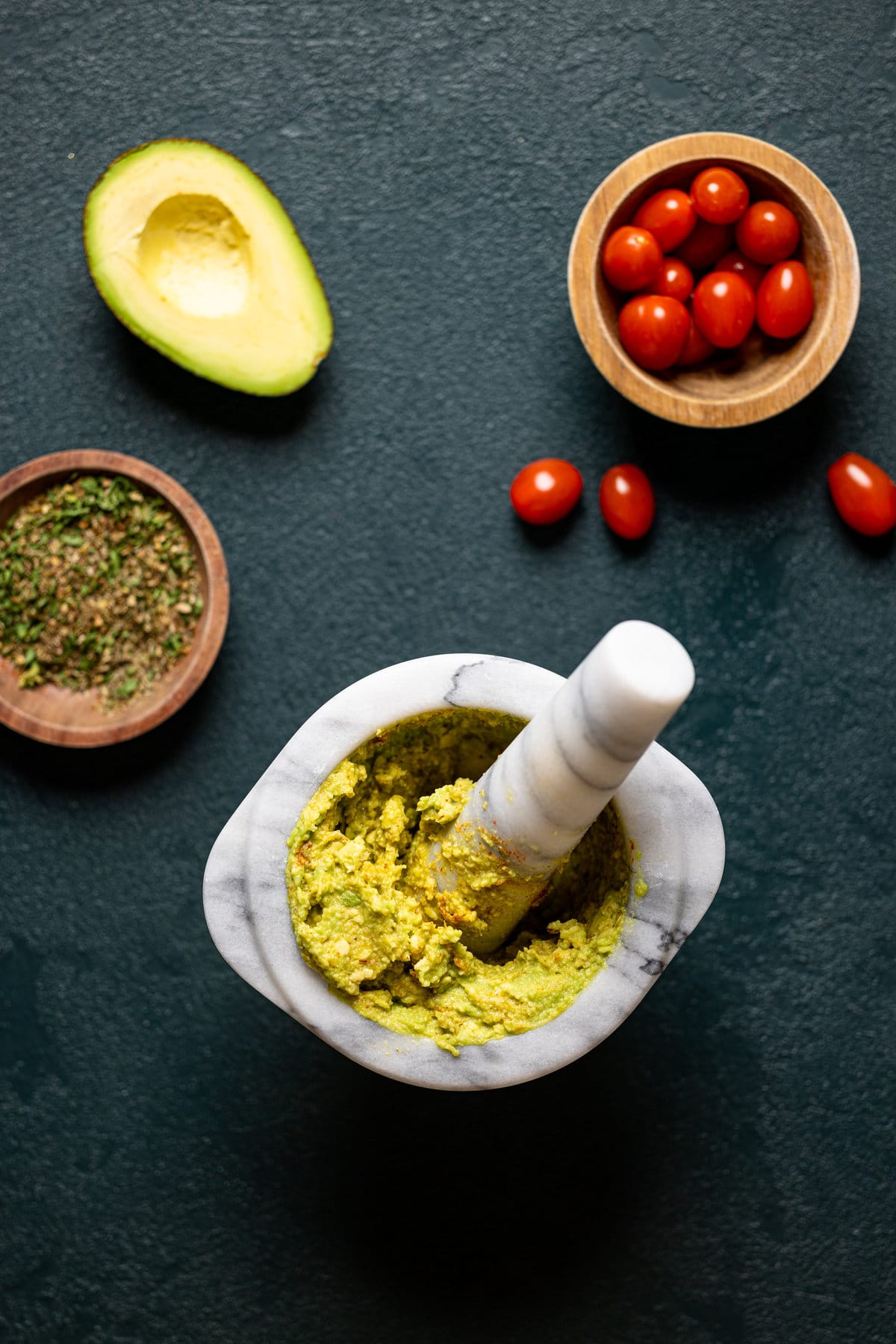 Mortar and pestle with an avocado mash next to tomatoes, half of an avocado, and seasonings