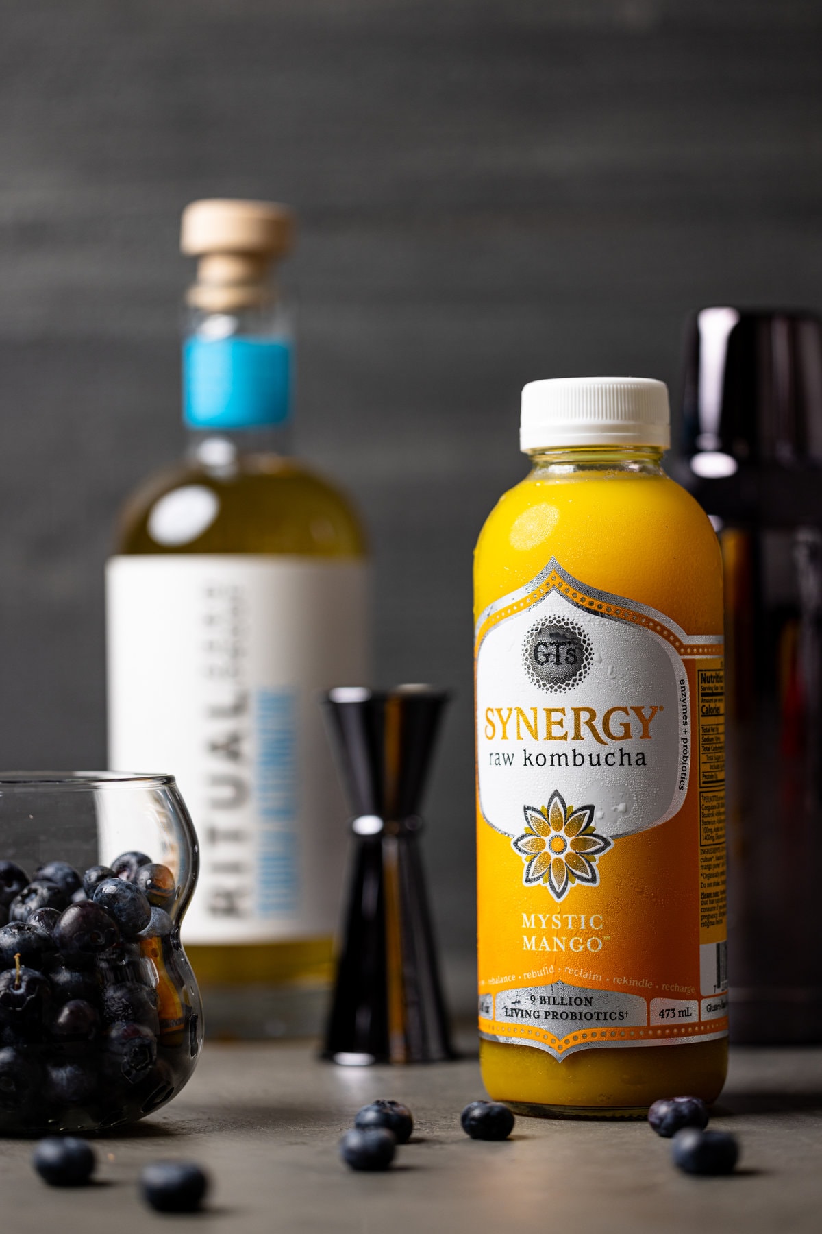 Mystic Mango flavored Synergy raw kombucha near a glass of blueberries