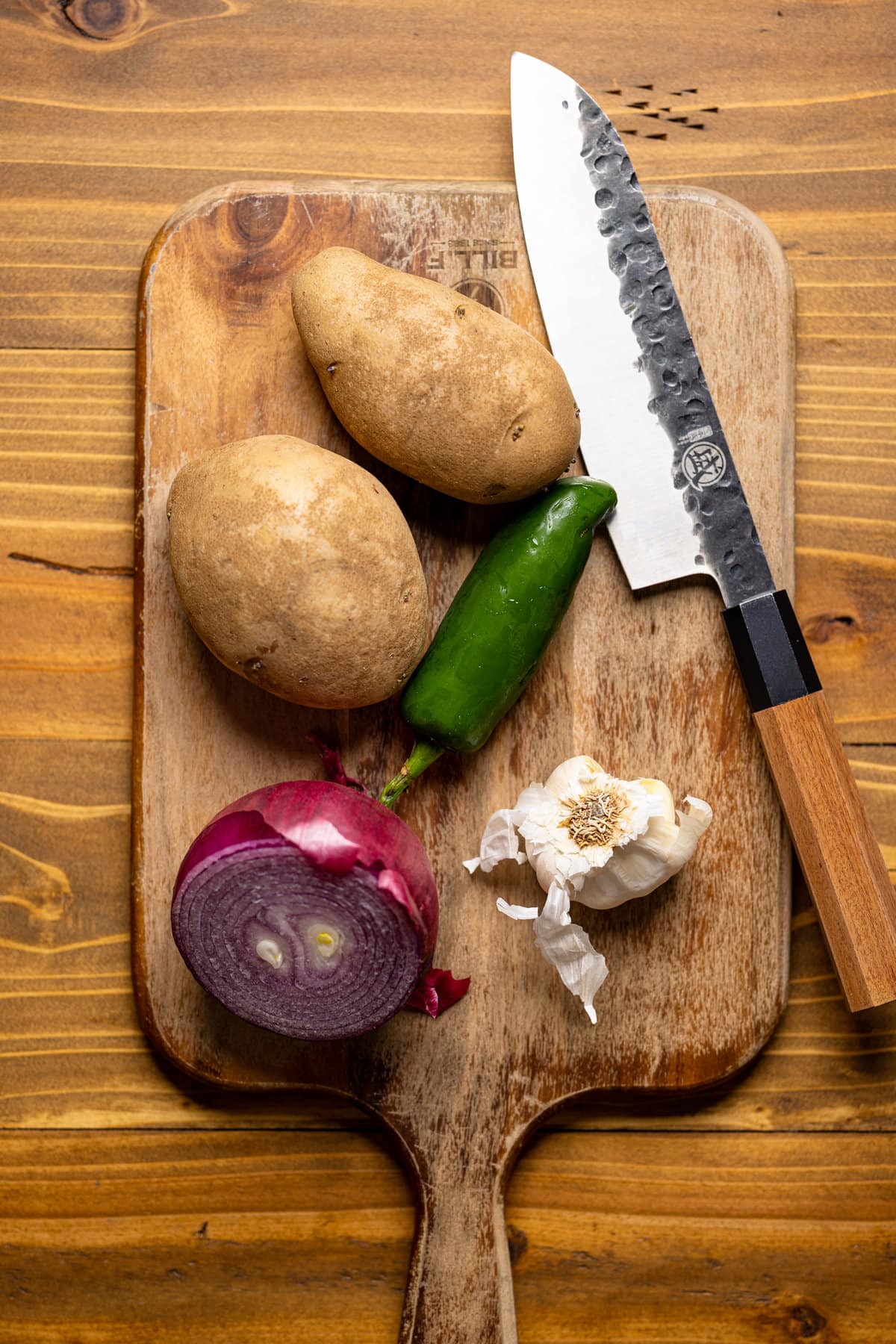 Potatoes, pepper, garlic, and red onion on a cutting board with a knife