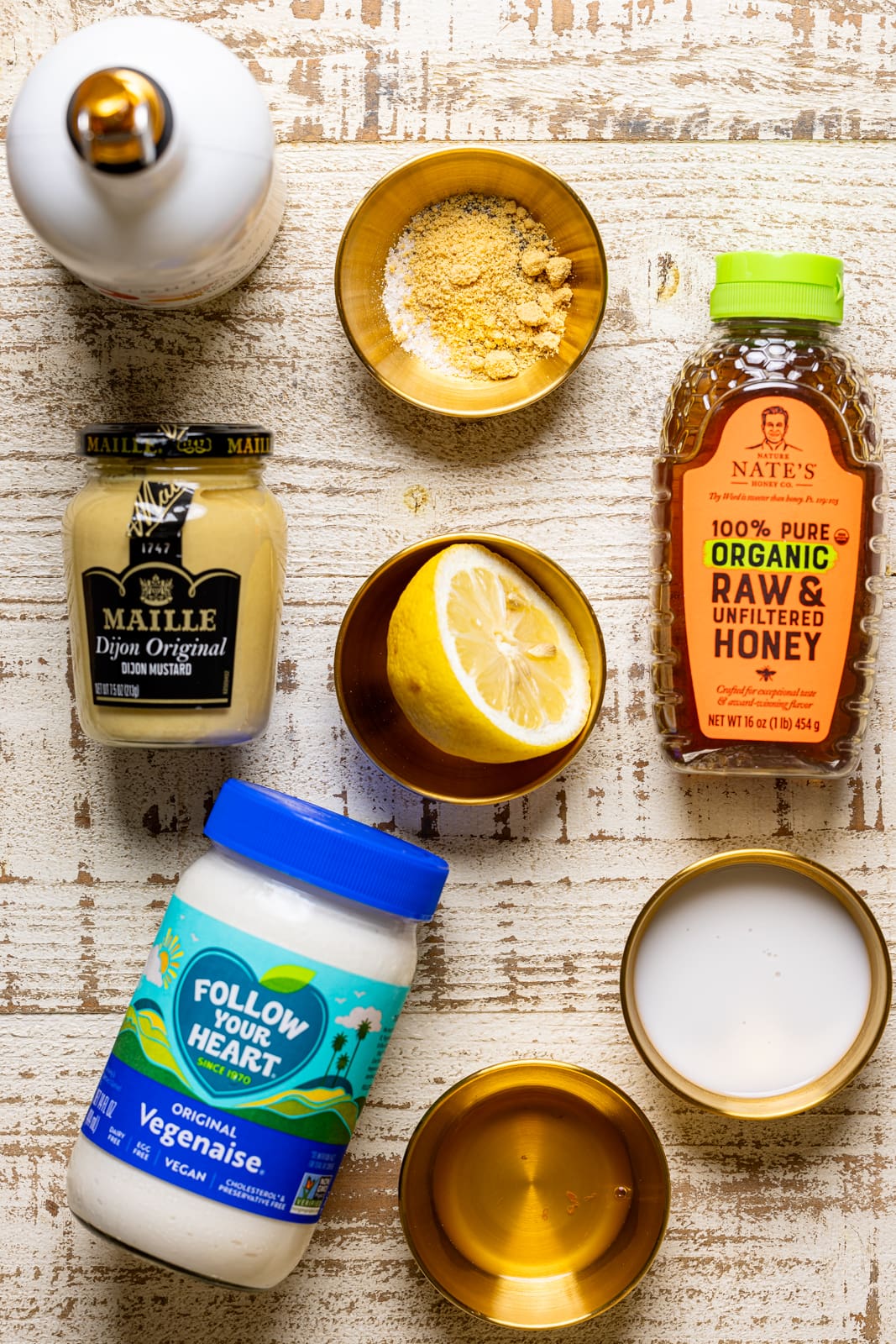 Ingredients on a white wood table including vegan mayonnaise, honey, lemon, dijon mustard, milk, seasoning and herbs, and olive oil.