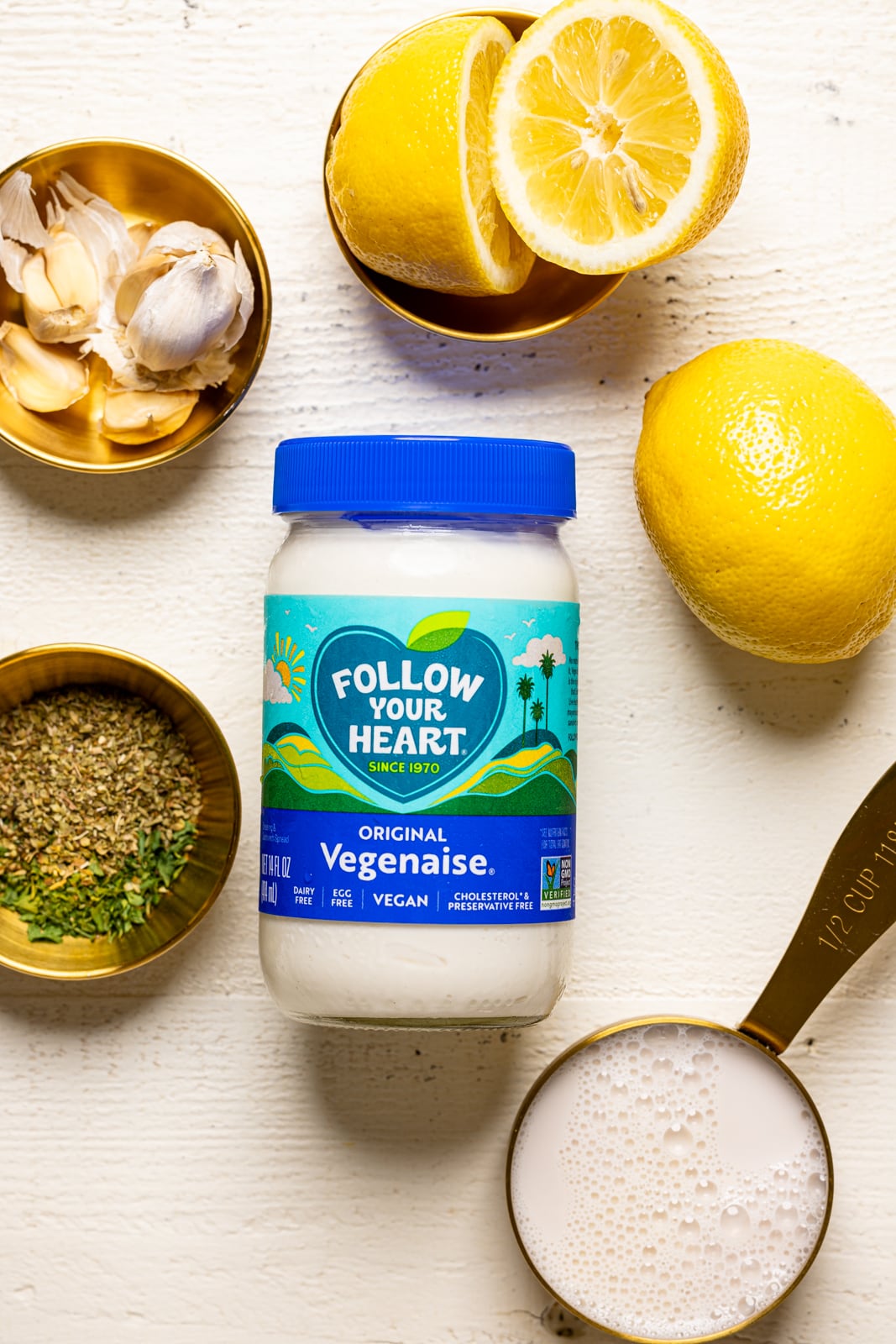 Dressing ingredients on a white table: vegan mayonnaise, garlic cloves, lemon, seasonings and herbs, and milk.