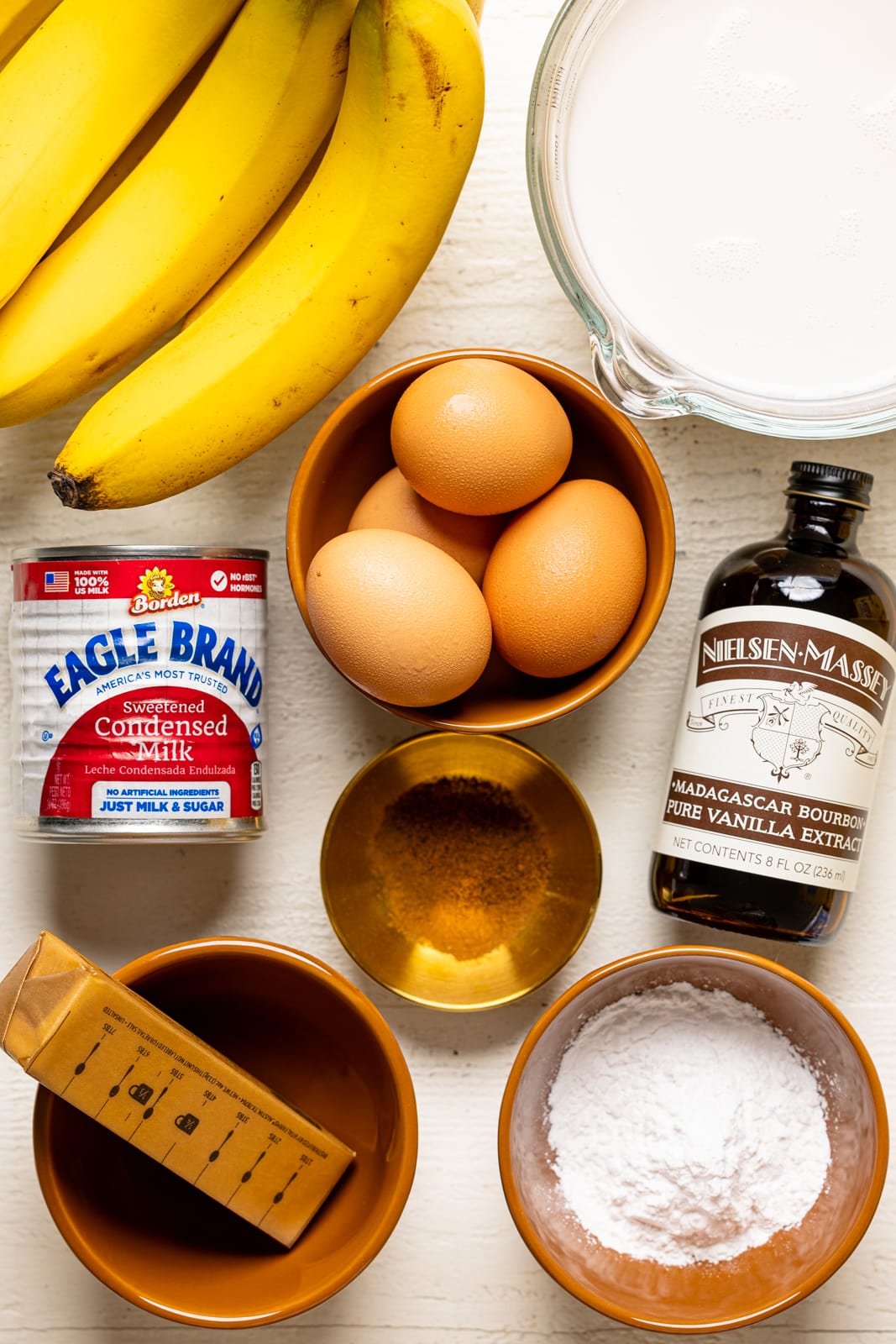 All ingredients on a white table that include condensed sweetened milk, bananas, vanilla extract, eggs, butter, milk, and cornstarch. 