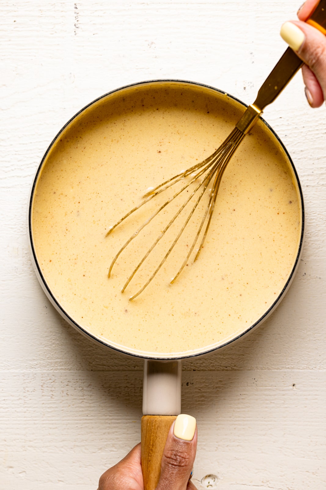 Homemade vanilla pudding in a pot with a gold whisk on a white table.