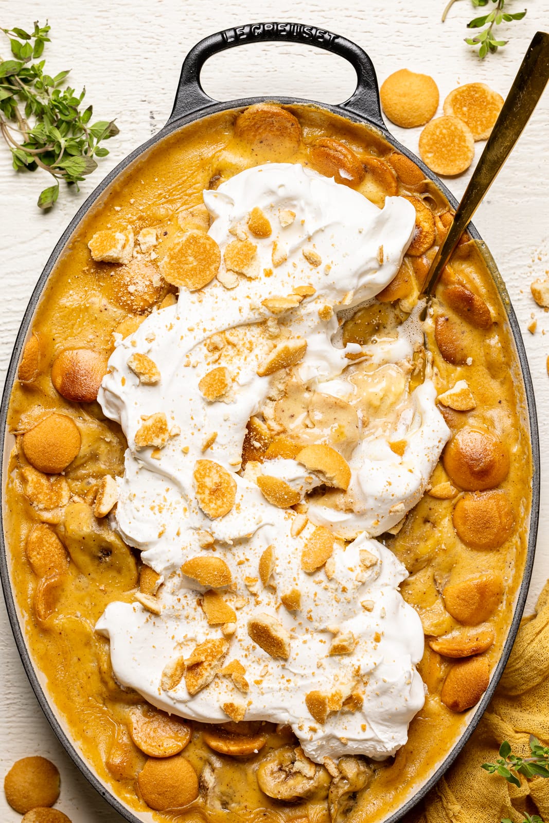 Baked banana pudding in a baking dish topped with whipped cream and a spoon on a white table. 