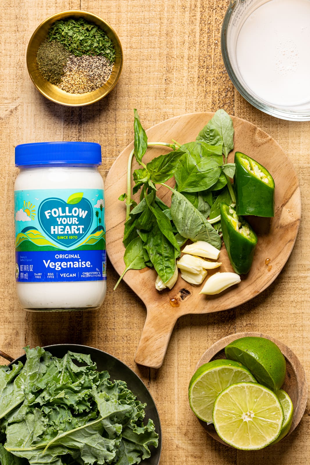 Ingredients on a brown wood table including vegan mayonnaise, jalapeños, garlic cloves, lime, seasonings and herbs, milk, basil, and kale.